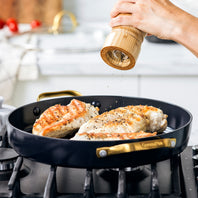 A person grinding pepper over chicken cooking in the GreenPan Reserve Ceramic Nonstick 11" Grill Pan in black with a gold-tone handle. 