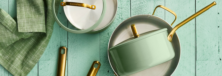 Three saucepans with gold handles, one green and two silver, are placed on a mint-green wooden surface next to a green cloth napkin.