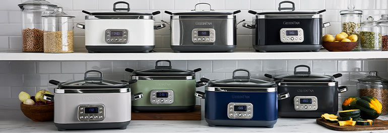 Eight slow cookers in various colors are displayed on a kitchen counter and shelf, surrounded by jars of grains, vegetables, and lemons, against a white tile backsplash.