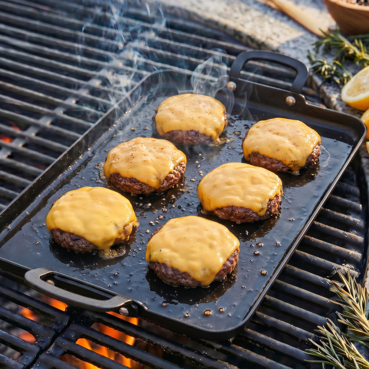 Six burger patties with melted cheese cooking on a rectangular carbon steel double burner griddle, smoke rising.