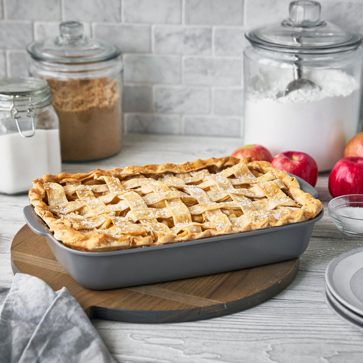 Lattice-topped pie in a rectangular GreenPan baking dish on a wooden board, with apples and sugar jars in the background.