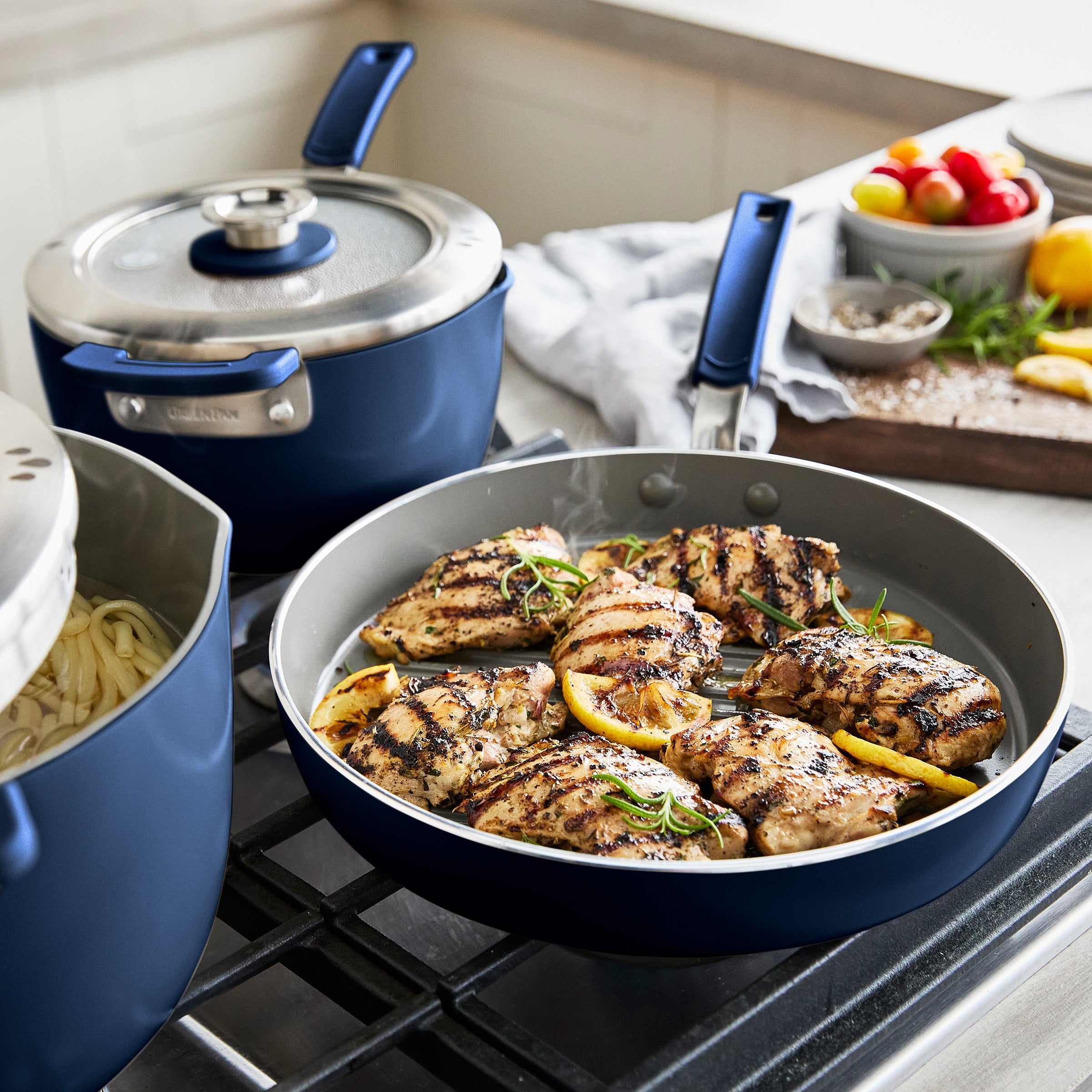 Cooked chicken with lemon and herbs in a frypan on a stove, with kitchenware and ingredients in the background