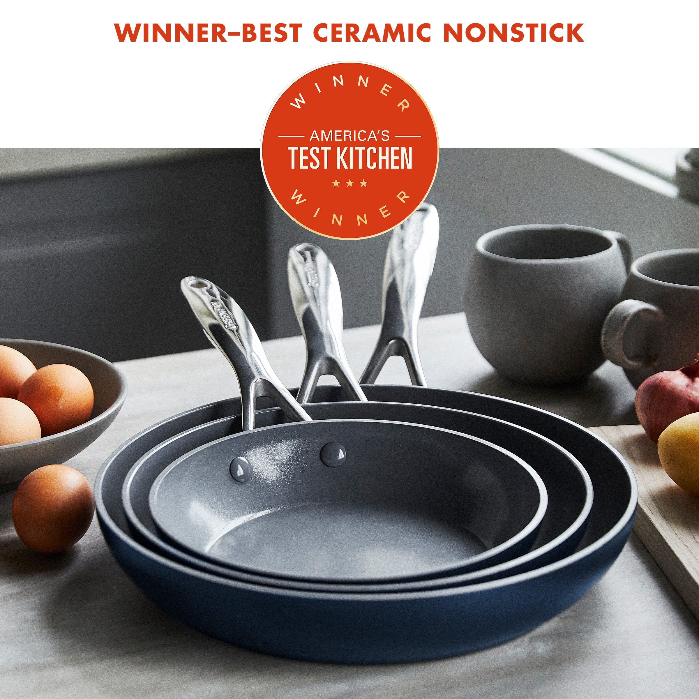 Set of three ceramic skillets stacked on a kitchen counter, with eggs and dishes nearby. Text reads: “Winner-Best Ceramic Nonstick.”.