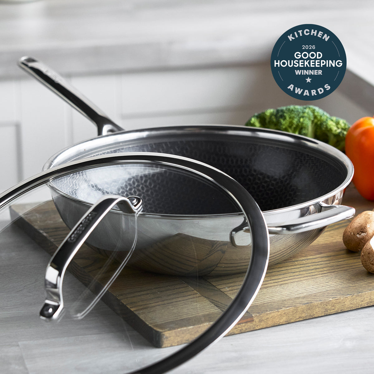 Gray ceramic 12-inch wok with glass lid, surrounded by assorted fresh vegetables on a kitchen counter.