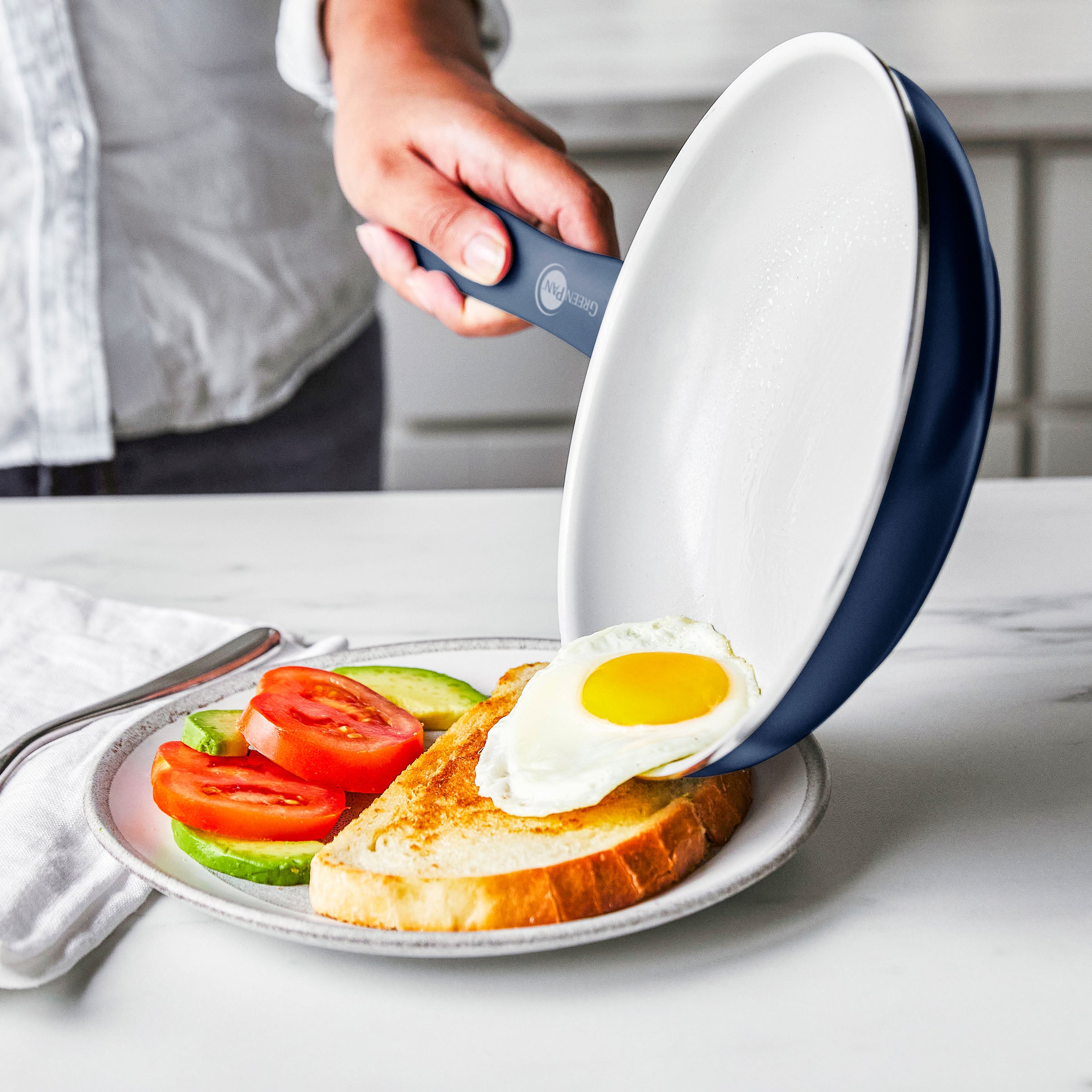 A hand slides a sunny-side-up egg from a ceramic nonstick pan onto toast with tomato and avocado on a white plate.
