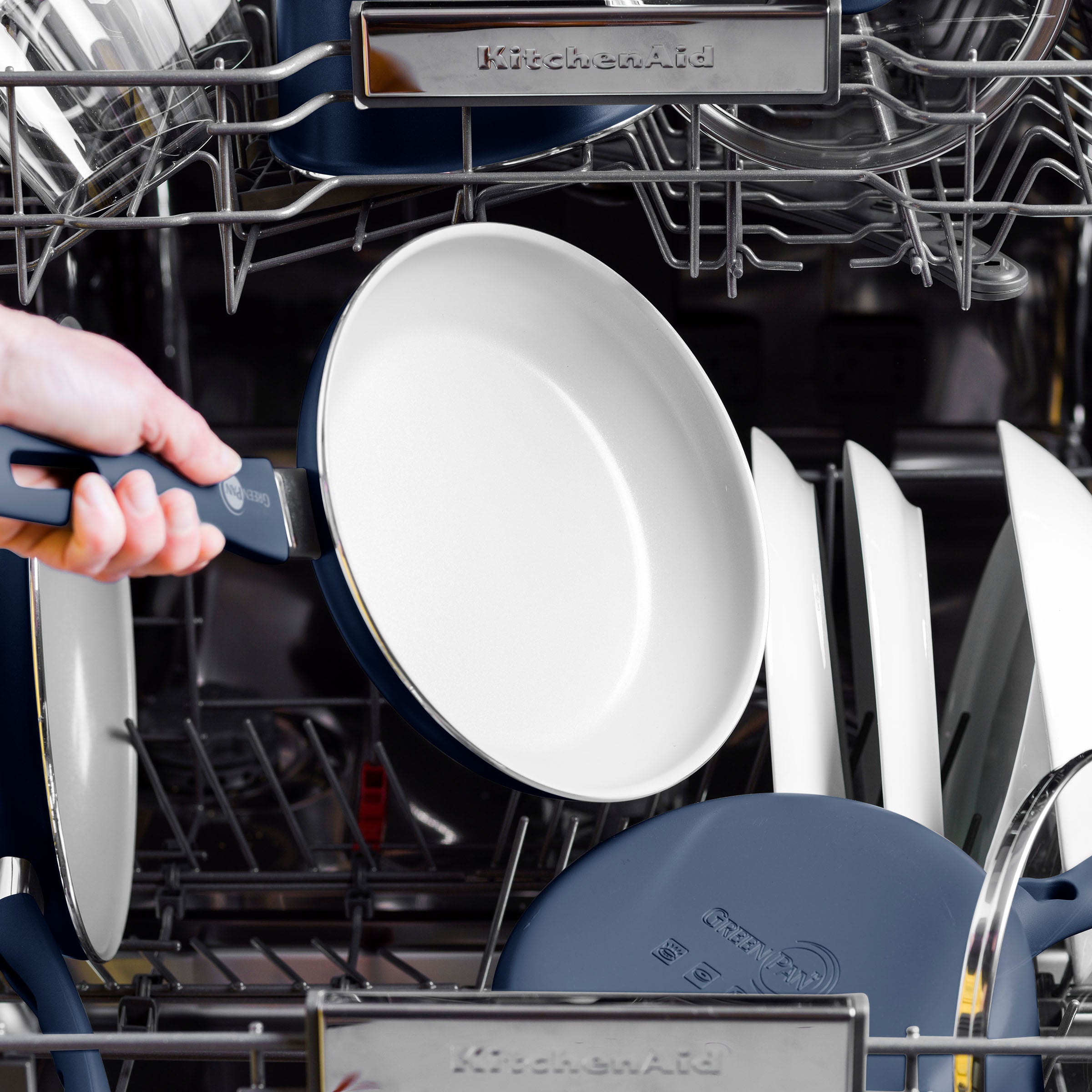 A hand loads a blue ceramic nonstick pan with a dark handle into a dishwasher filled with assorted dishes.