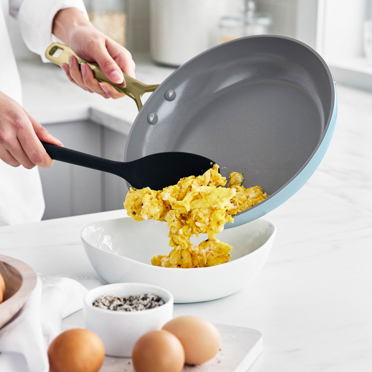 Scrambled eggs being served from a Glacier-colored ceramic nonstick pan into a white bowl on a kitchen counter.