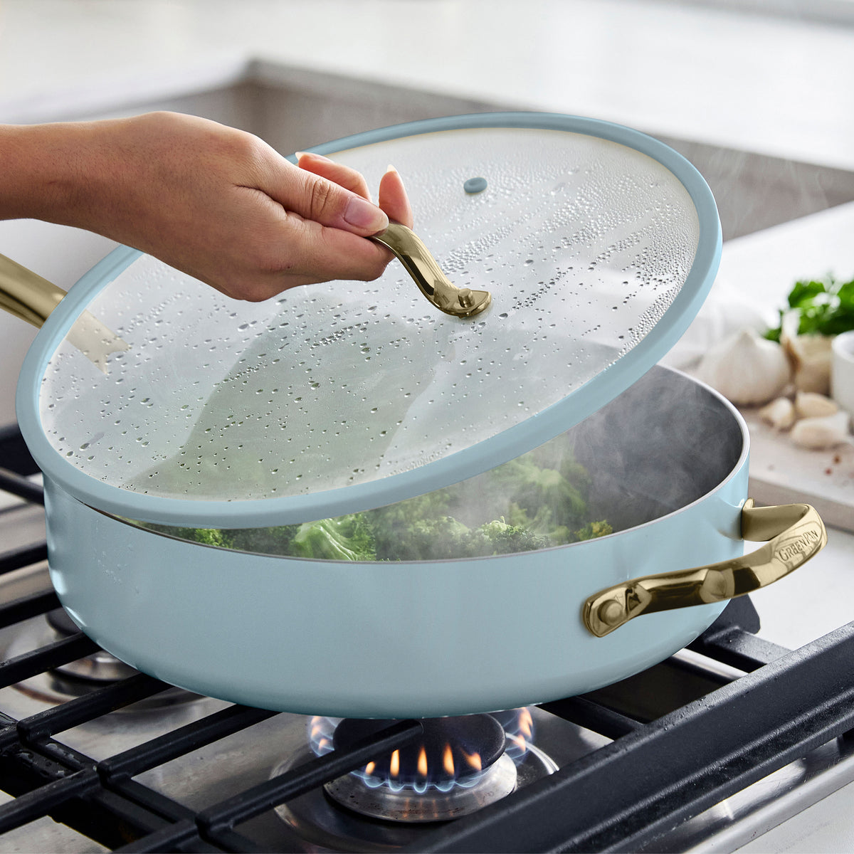 Hand lifting lid from a steaming ceramic nonstick pan in Glacier color with broccoli cooking on a gas stove.