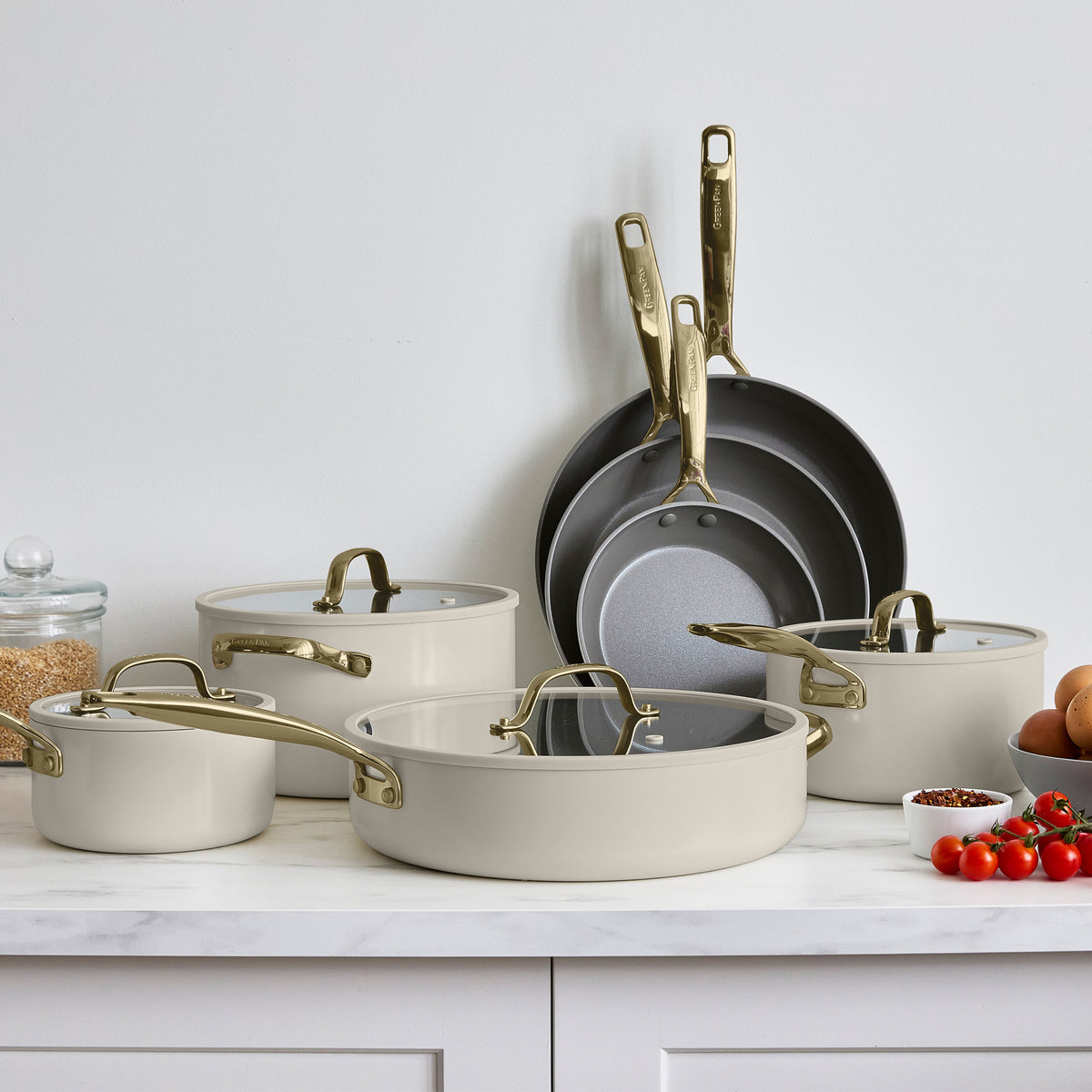 Taupe ceramic nonstick 11-piece cookware set with gold handles arranged on a white countertop next to fresh ingredients.