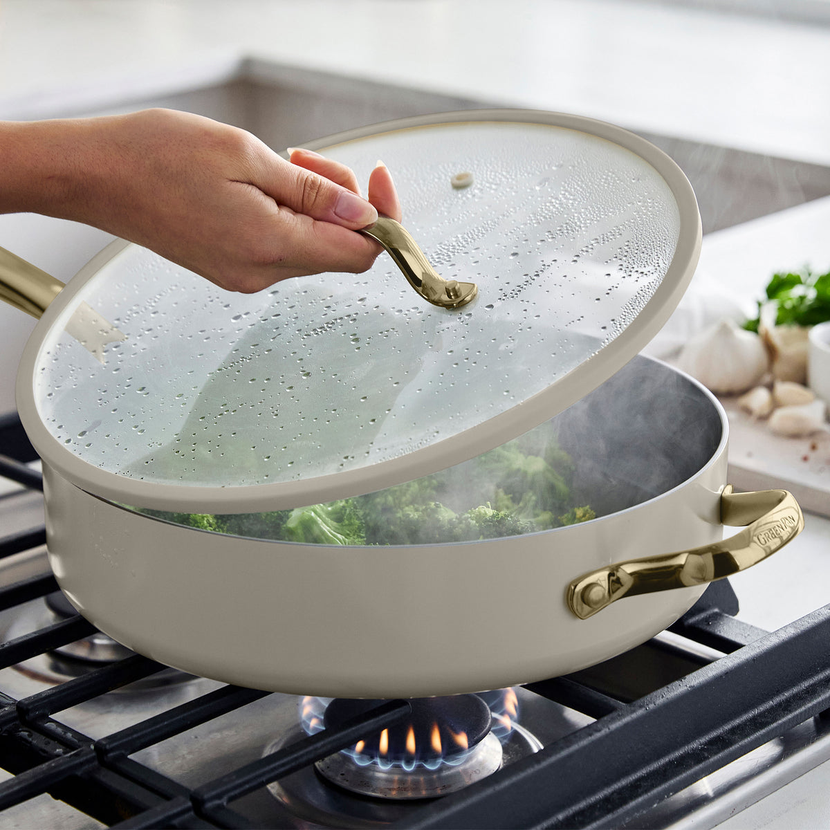 Hand lifting glass lid from a taupe ceramic nonstick pan with steamed broccoli on a gas stove.