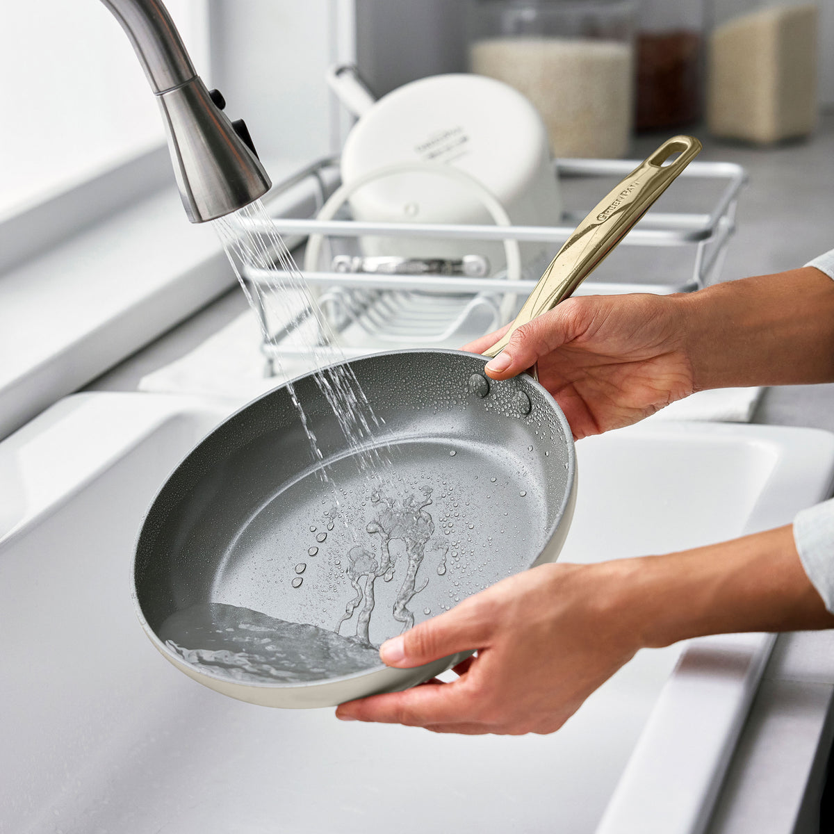Person rinsing a taupe ceramic nonstick pan from an 11-piece cookware set under water at a kitchen sink.