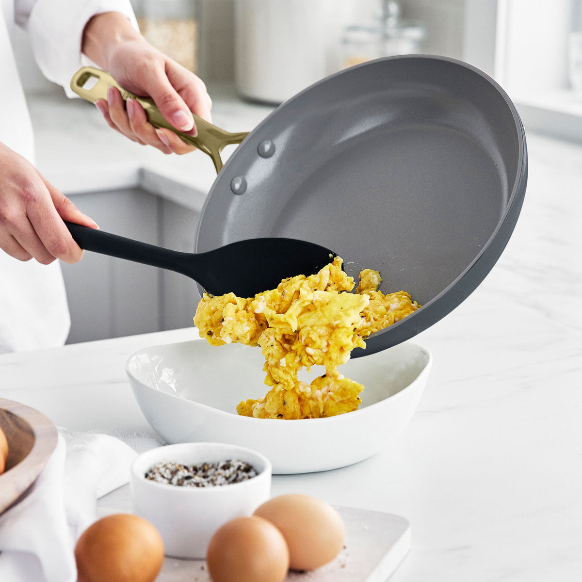 Scrambled eggs being served from a gray ceramic nonstick pan into a bowl, with whole eggs and pepper on the counter.