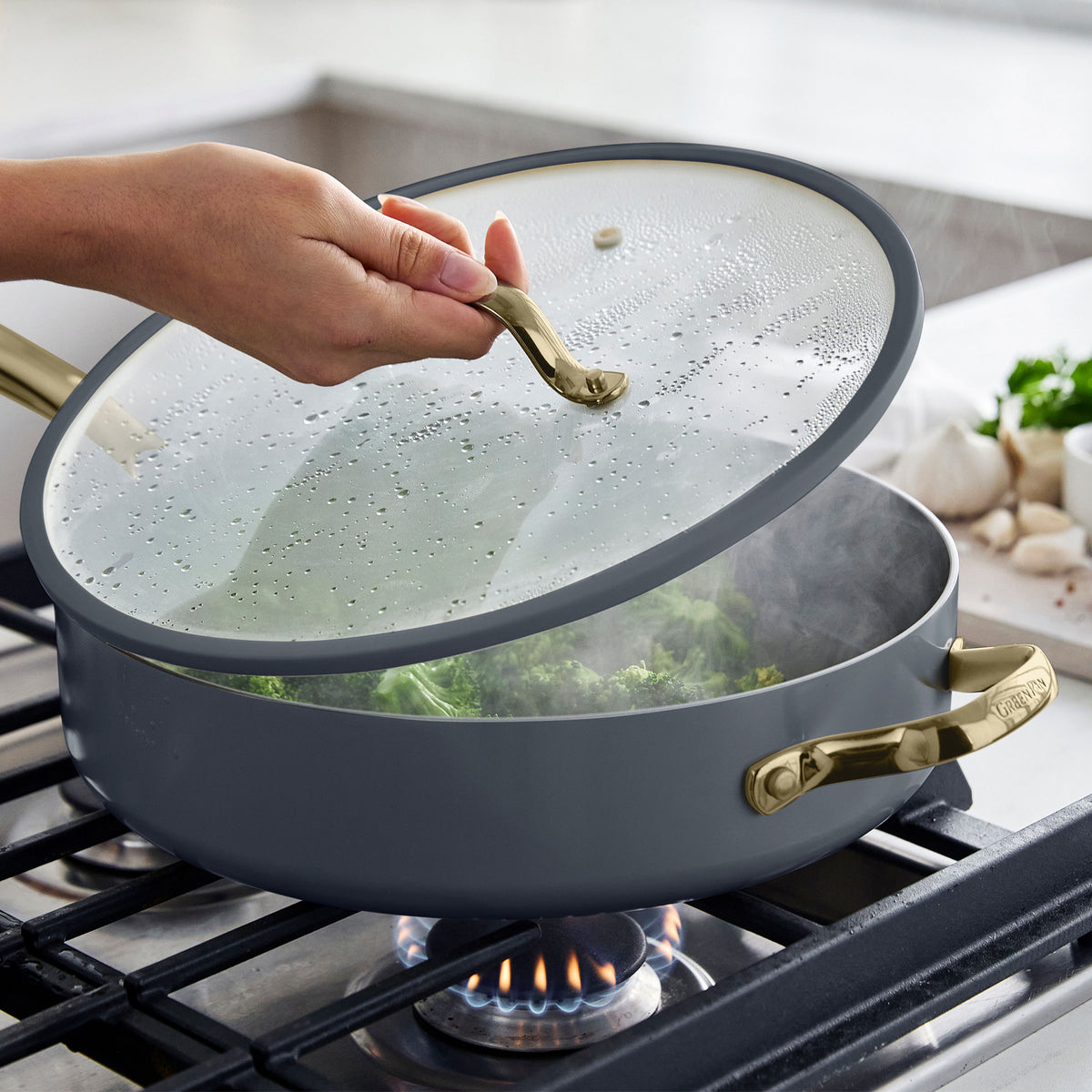 Hand lifting glass lid of a slate ceramic nonstick pan cooking vegetables on a gas stove with brass handles.
