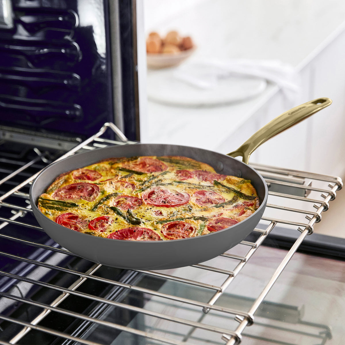 Frittata with tomatoes and greens in a ceramic nonstick skillet on a stove, oven rack and counter visible in the background.