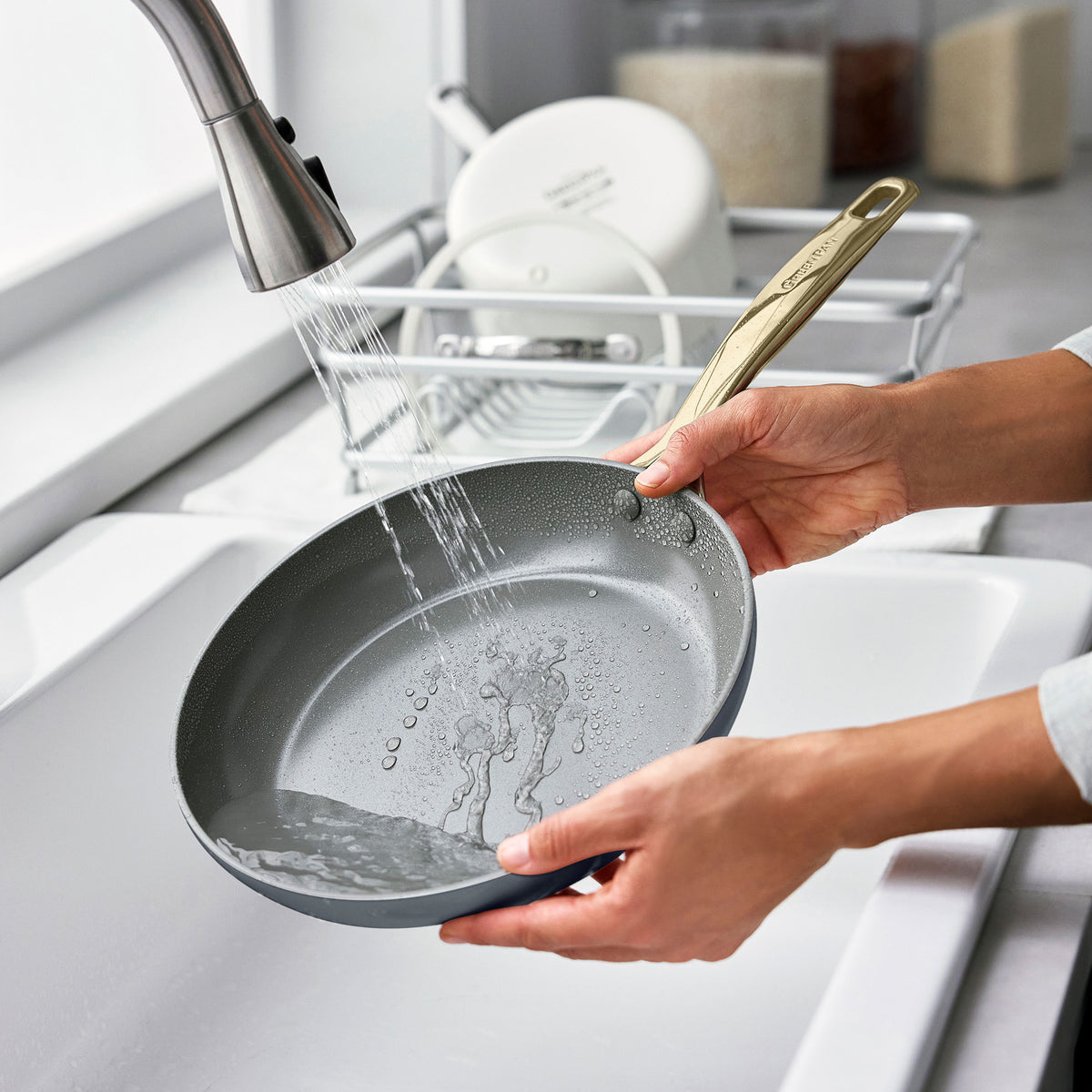 A hand rinses a gray ceramic nonstick pan from an 11-piece set under a kitchen faucet beside a dish rack with plates and bowls.