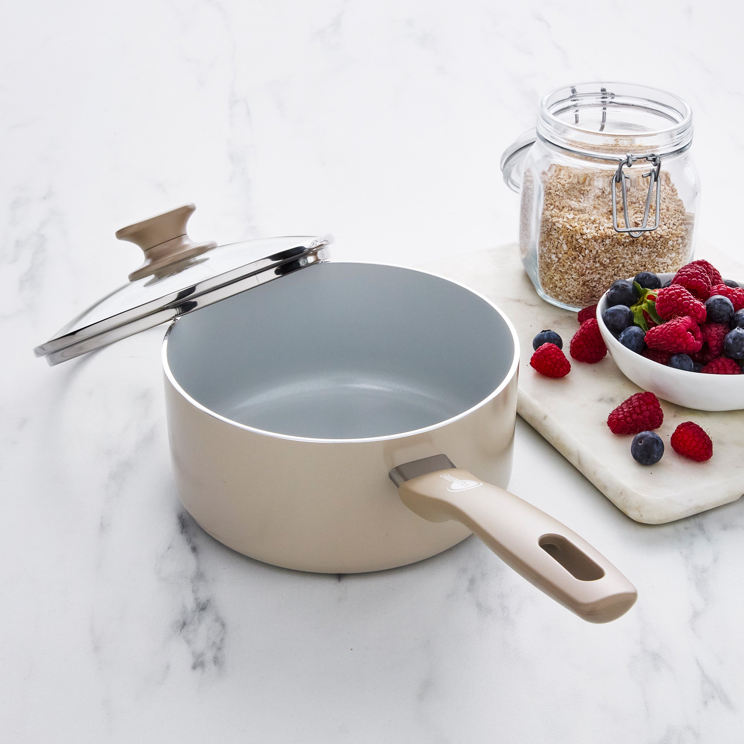 Taupe ceramic nonstick saucepan with lid beside oats and berries on a marble countertop.