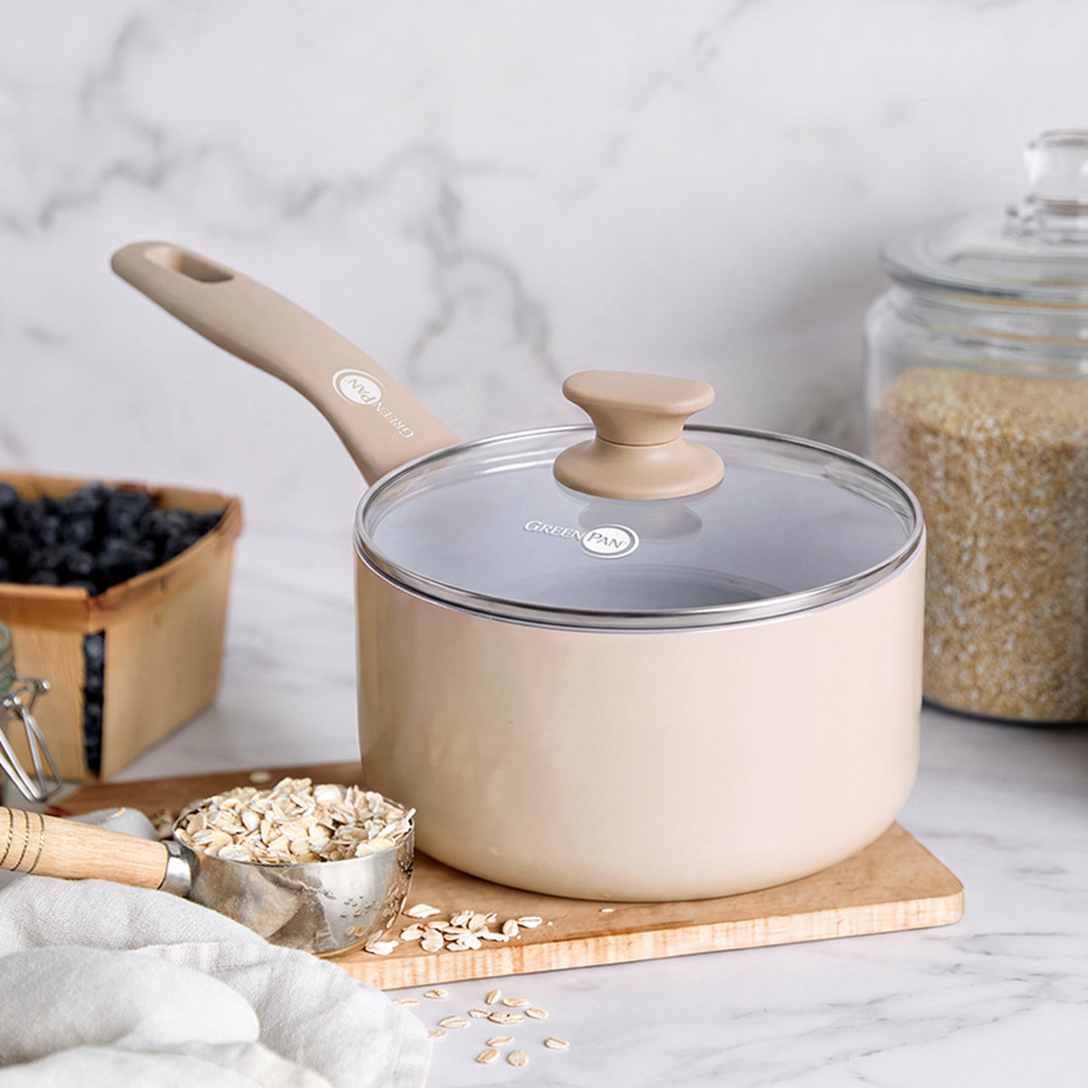 Taupe ceramic nonstick saucepan with lid on wood surface, surrounded by oats, blueberries, and grains on a marble background.