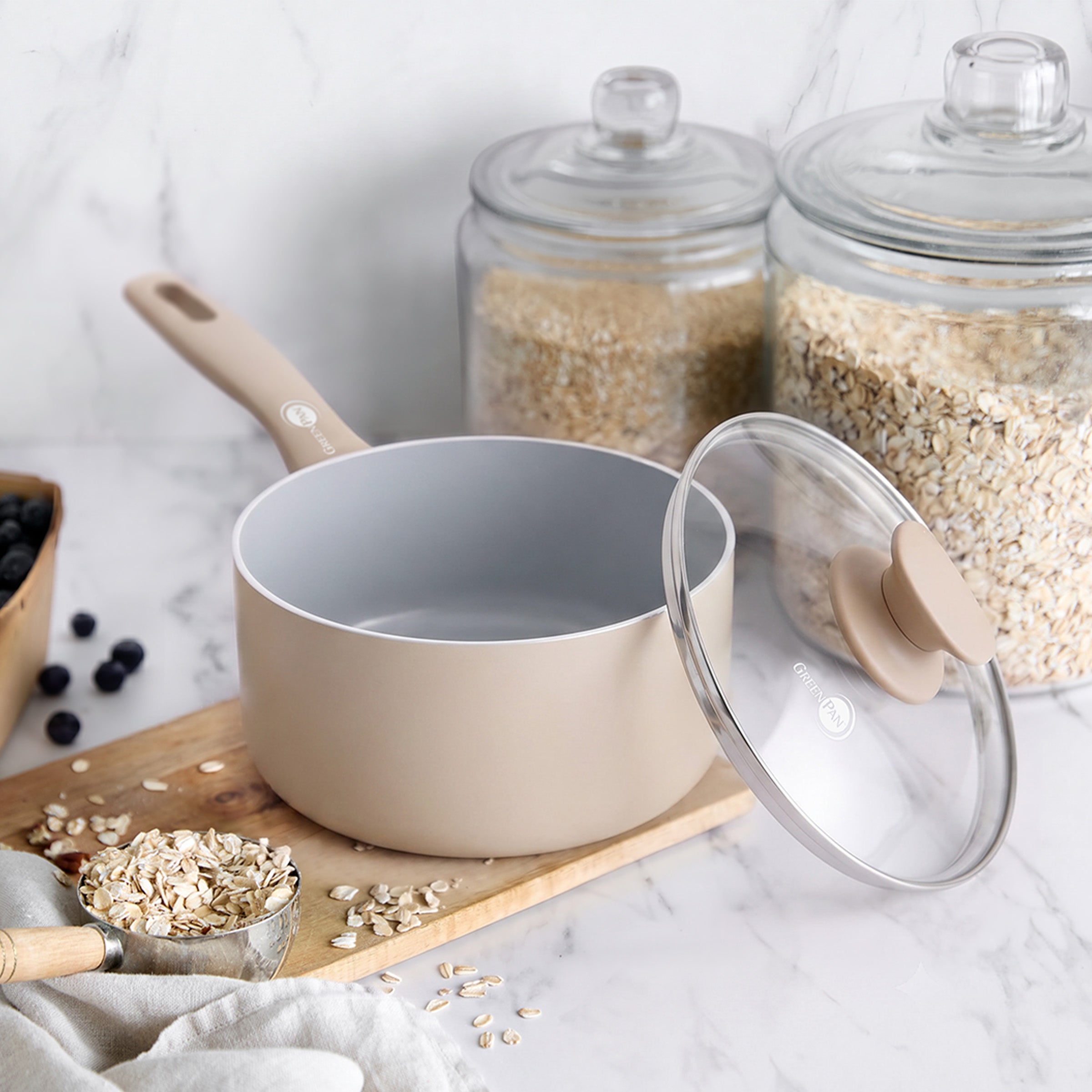 Taupe ceramic nonstick saucepan with lid on a board, next to glass jars of oats and a bowl of blueberries.