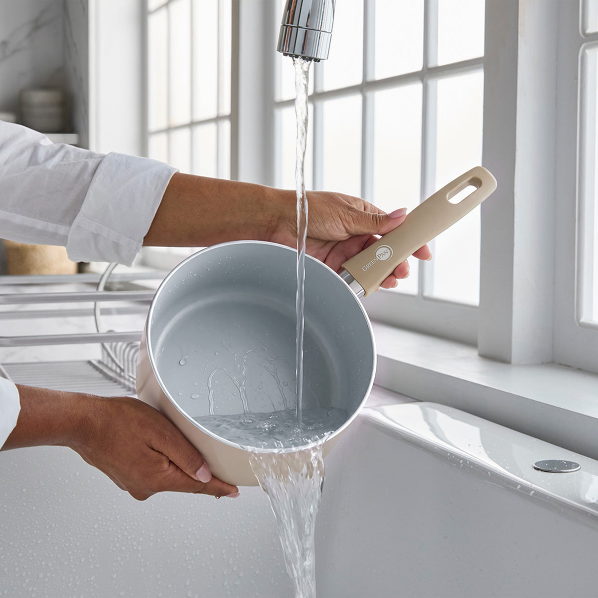Rinsing a taupe ceramic nonstick saucepan with lid at a kitchen sink near a window.