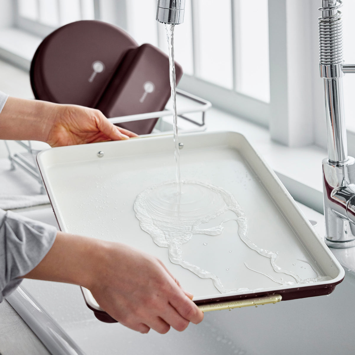 Hands rinsing a Bordeaux Reserve Bakeware 13″ x 18″ half sheet pan under a kitchen faucet.
