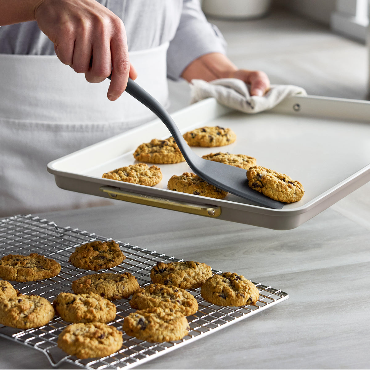 A person uses a grey spatula to move a cookie from a latte-colored Reserve Bakeware 13″ x 18″ Half Sheet Pan to a cooling rack.