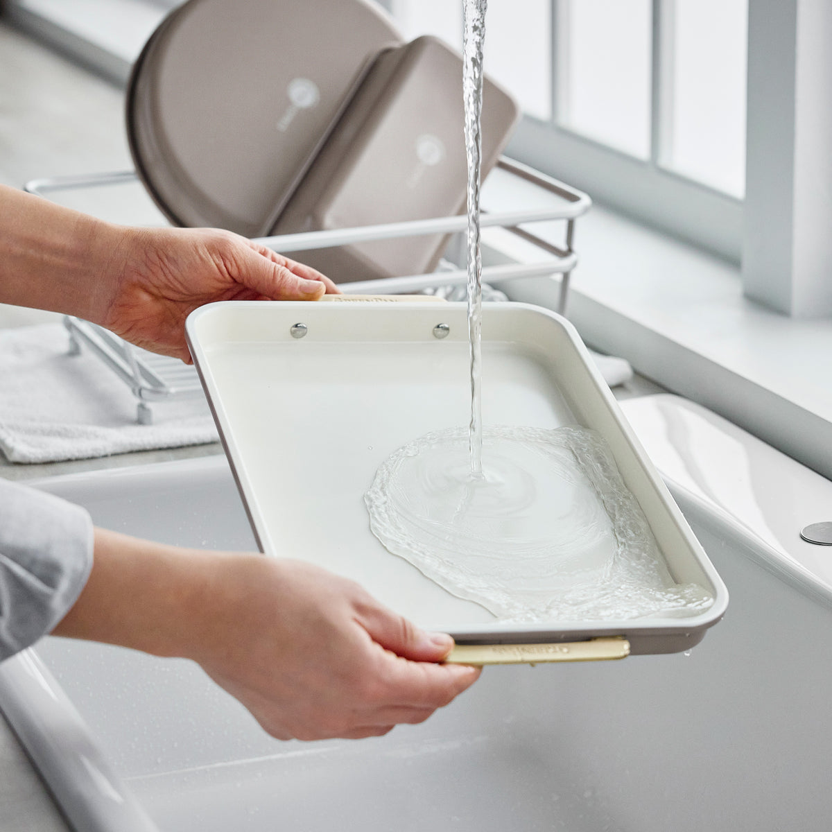 Hands rinsing a latte-colored, heavy-gauge aluminized steel baking pan from the Reserve Bakeware 7-piece set under a kitchen faucet.