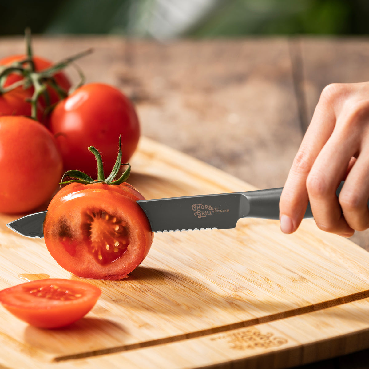 Hand slicing a red tomato with stainless steel knife from 2-piece set; whole tomatoes and extra blades on cutting board.