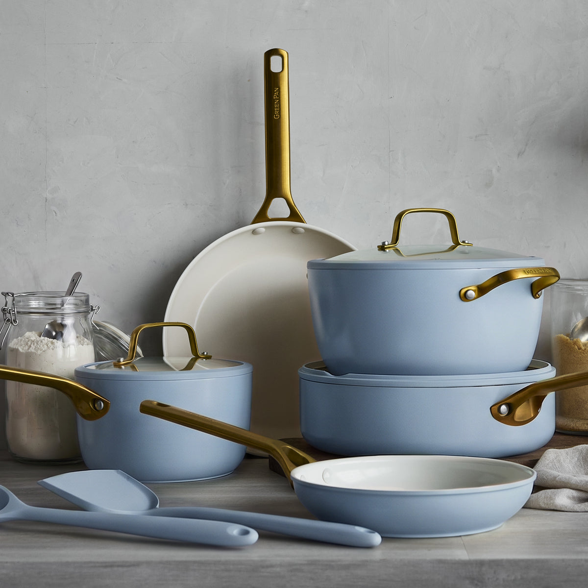 10-piece ceramic cookware set in light blue with gold handles, arranged on a kitchen counter beside flour and sugar jars.