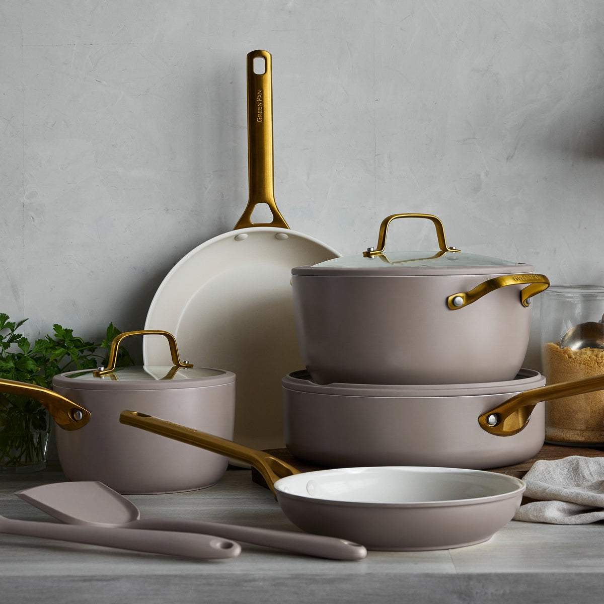 10-piece ceramic cookware set in latte with gold handles, spatulas, and fresh herbs displayed on a kitchen counter.