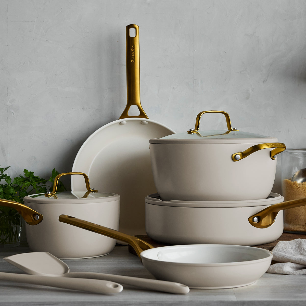 10-piece macadamia ceramic cookware set with gold handles, arranged on a kitchen counter next to utensils.