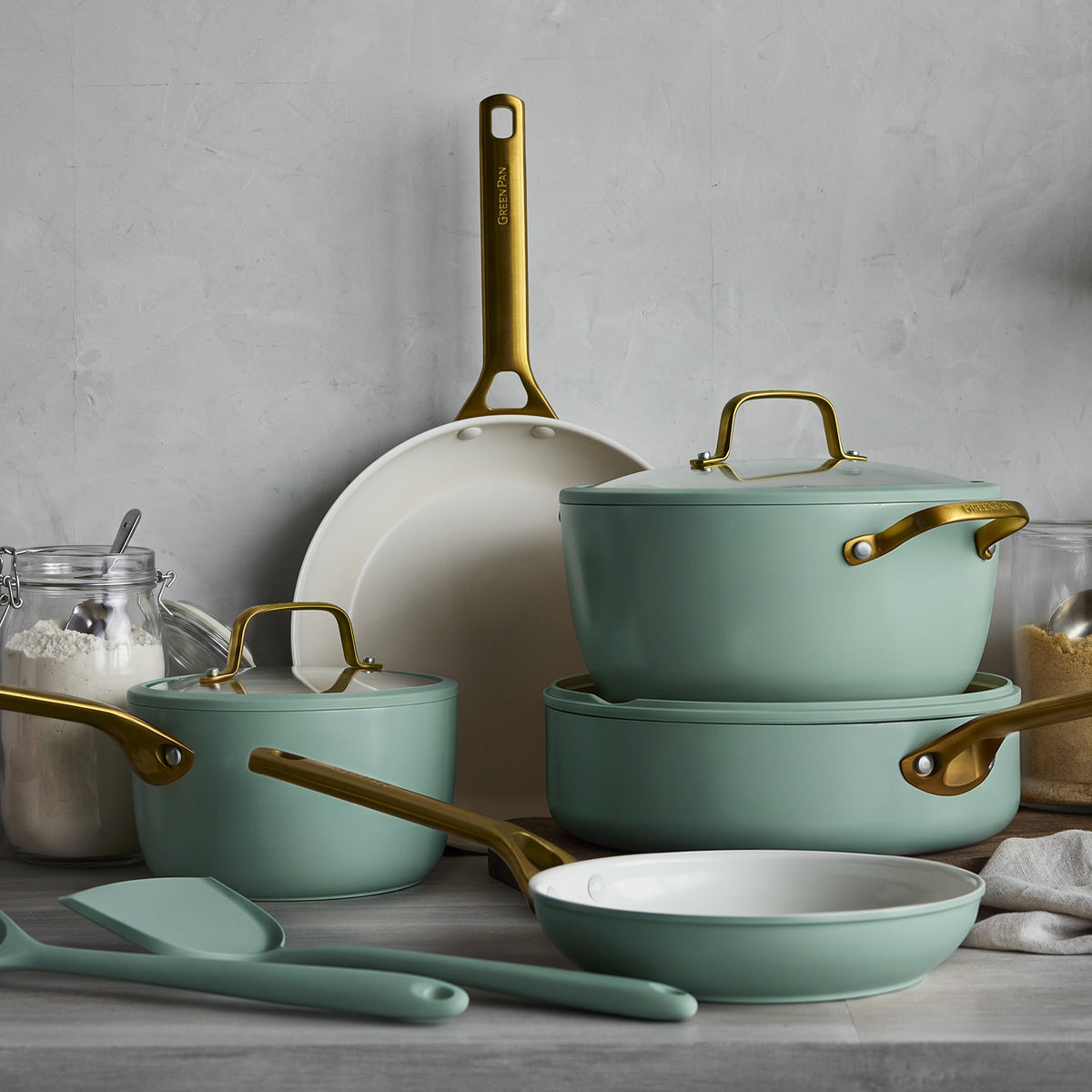 Green stackable pots and pans with gold handles on a kitchen counter near jars of flour and sugar.