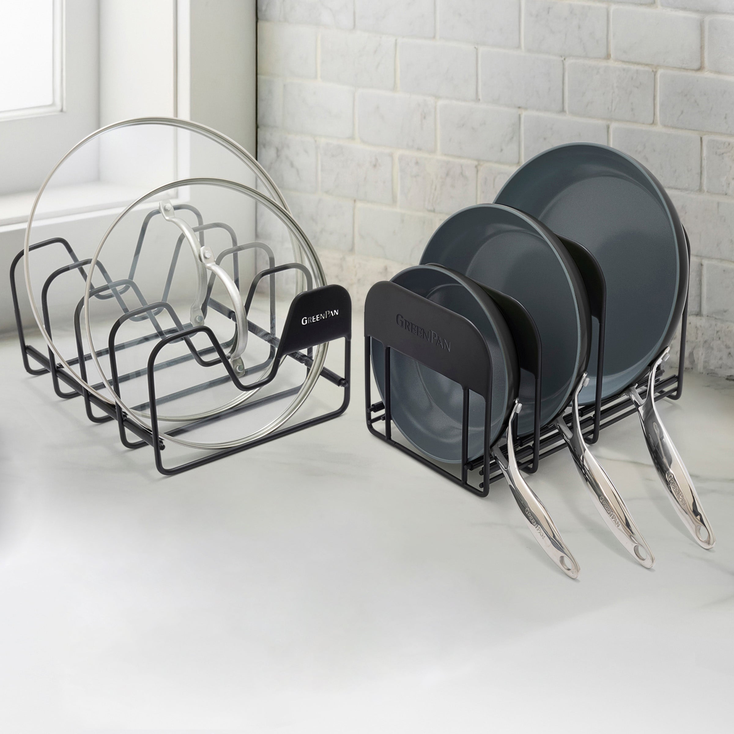 Two black racks holding glass pot lids and gray frypans set on a white kitchen counter near a window.