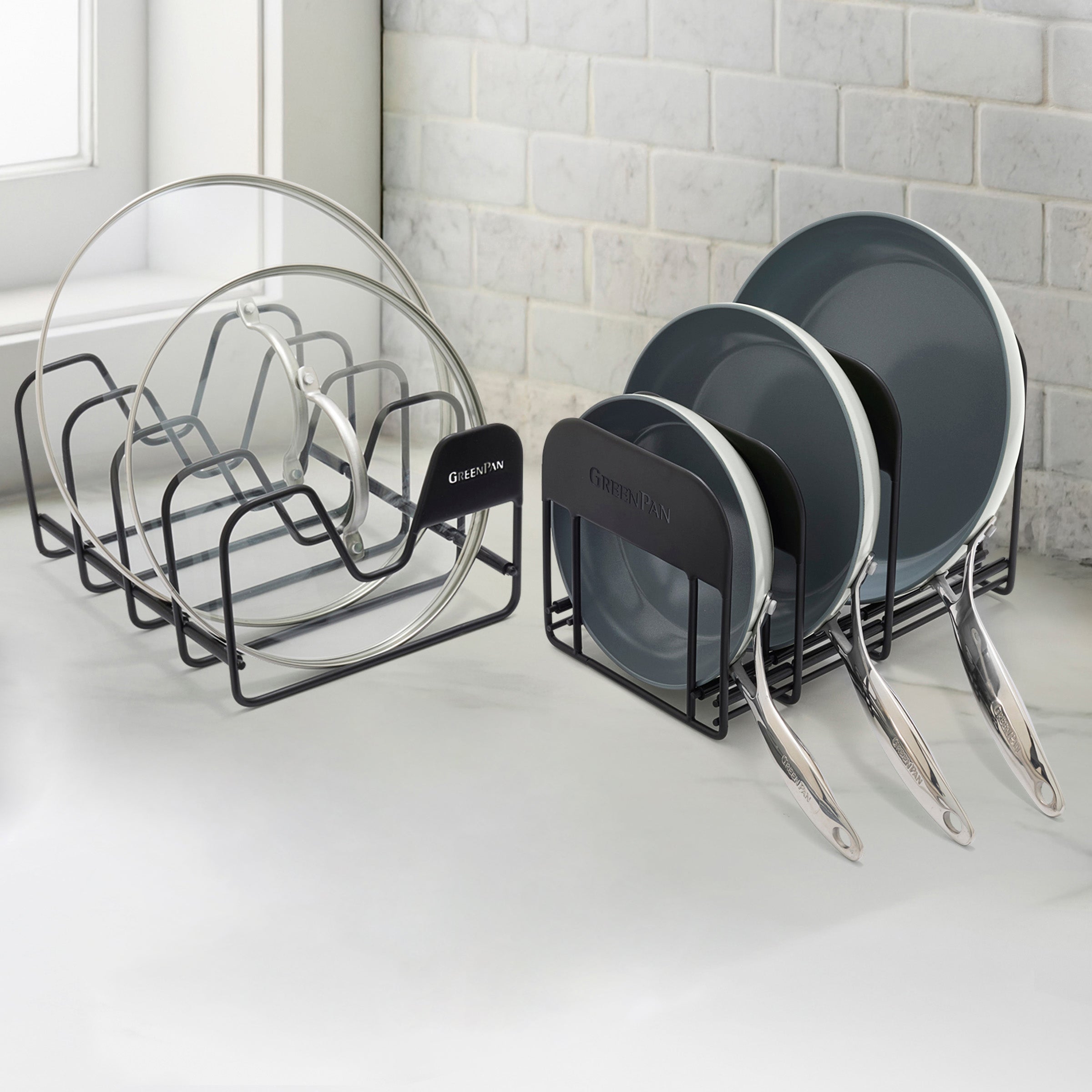 Two black racks display cream-colored frypans upright on a white kitchen counter in front of a tiled wall.