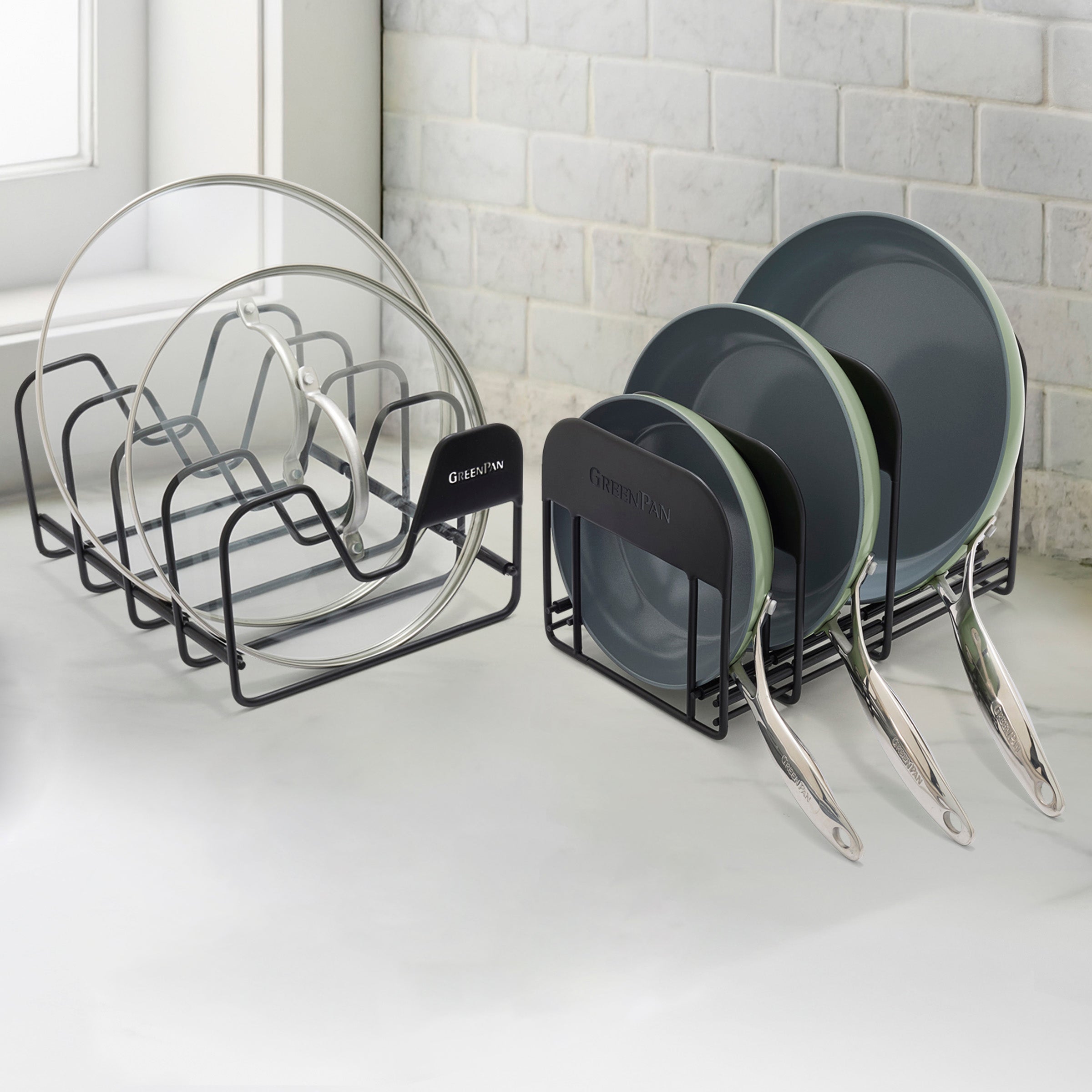 Green Valencia Pro frypan set with glass lids on a white countertop, held by black racks, with a tiled wall in the background.