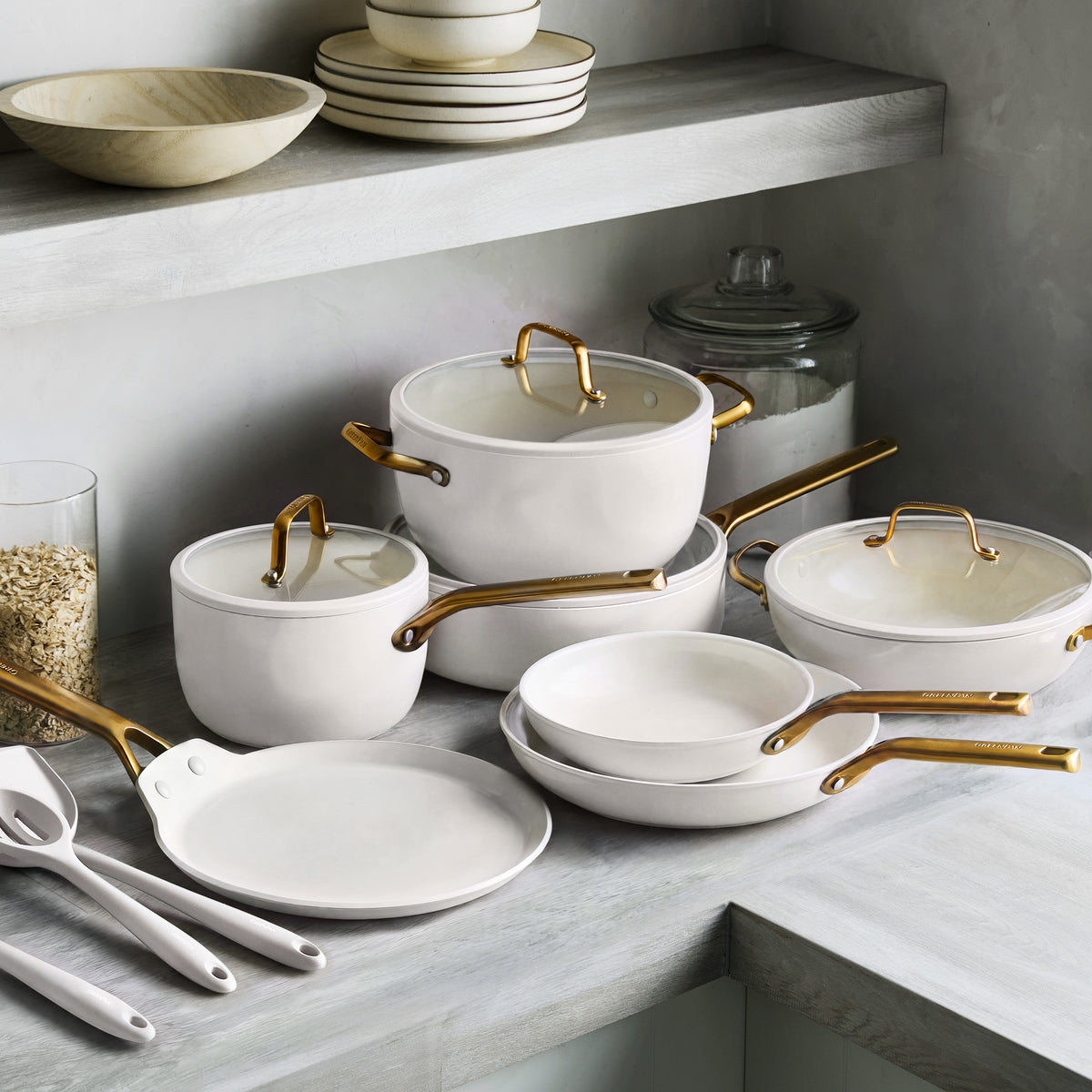 Cream ceramic nonstick cookware set with gold handles, 14 pieces, arranged on a kitchen counter near dishes and a jar.