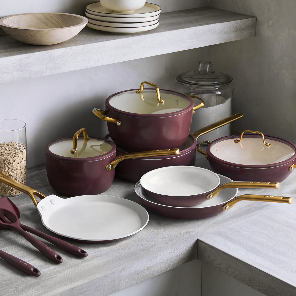 Bordeaux ceramic nonstick cookware set with gold handles, 14 pieces arranged on a gray counter alongside bowls and utensils.