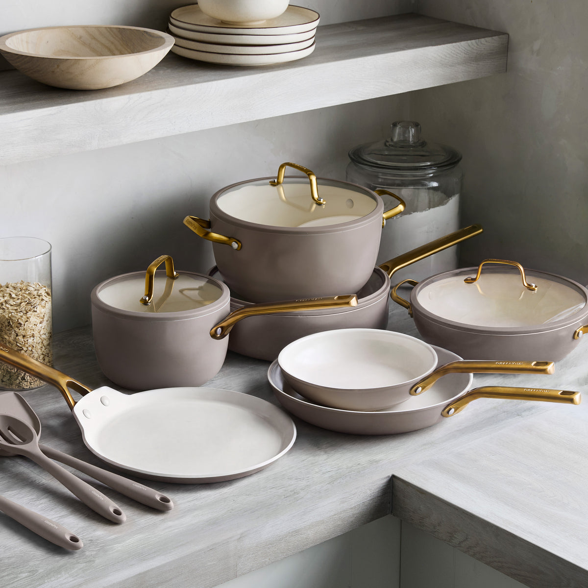 Latte-colored ceramic cookware set with gold handles, stackable pots, pans, and utensils arranged on a gray countertop.