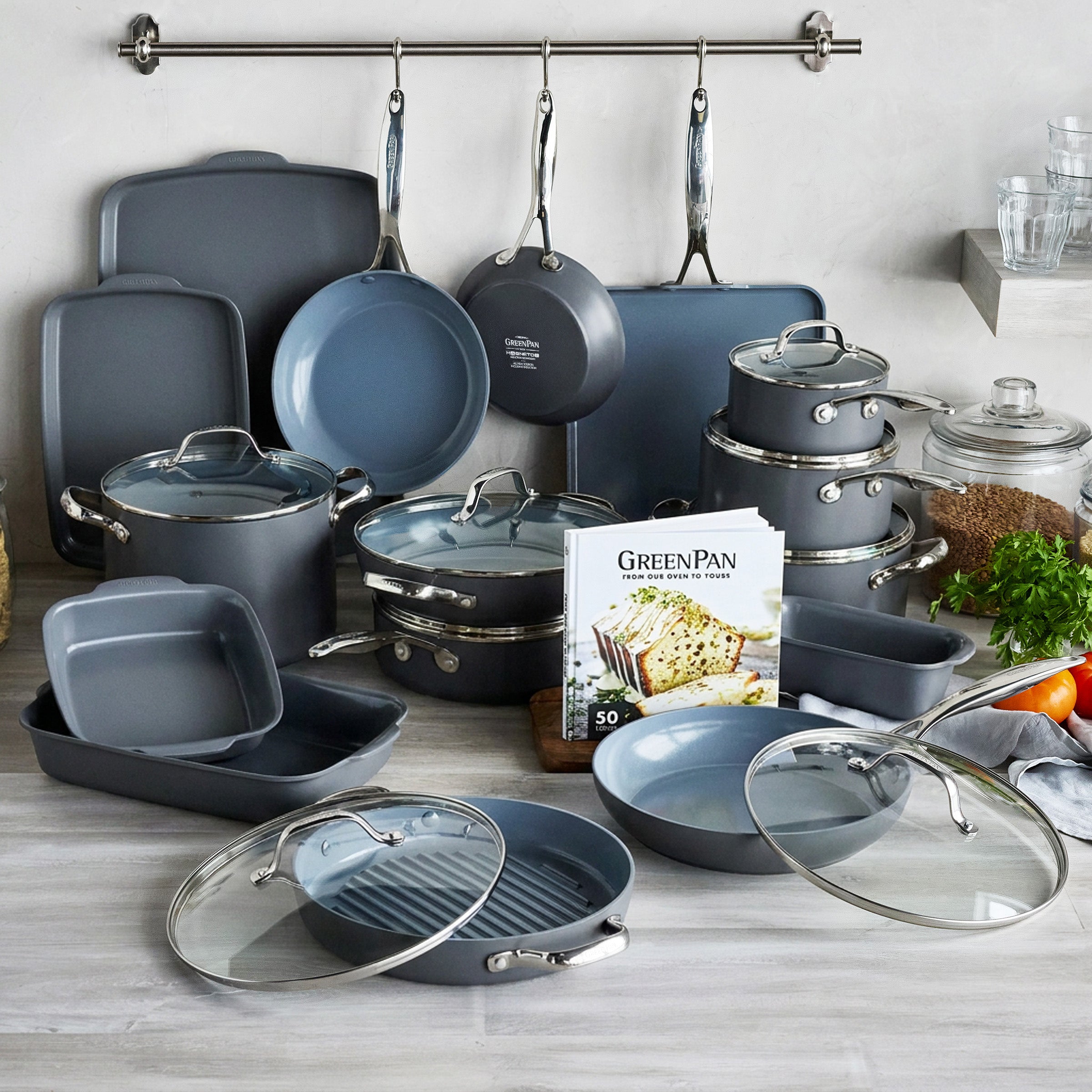 Gray 24-piece cookware and bakeware set with pots, pans, lids, and a cookbook arranged on a kitchen counter.