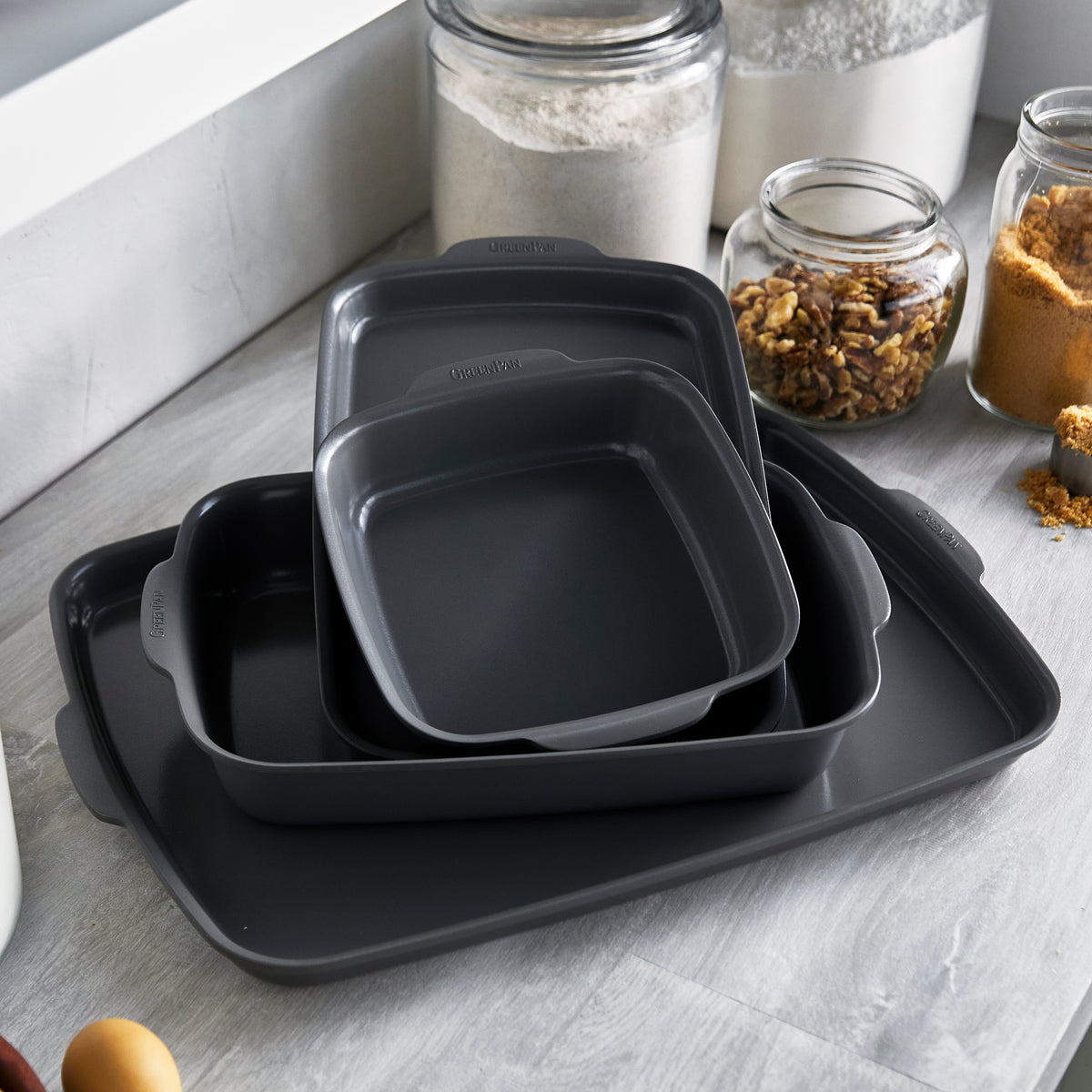 Set of four black ceramic baking dishes nested together on a countertop, with jars of flour, nuts, and brown sugar nearby.