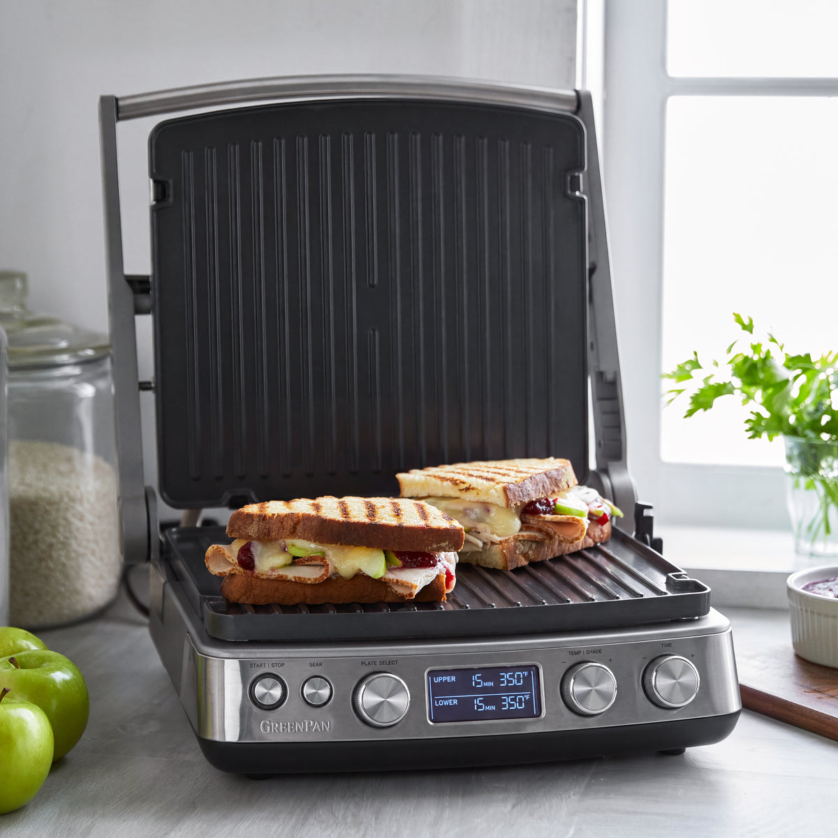 Two apple paninis grilling on a stainless steel Elite Multi Grill  Griddle in a sunlit kitchen.