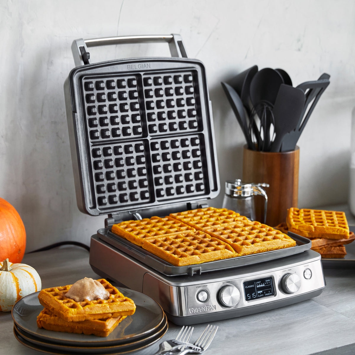 4-square ceramic waffle maker on a counter with pumpkin waffles, utensils, and small pumpkins nearby.