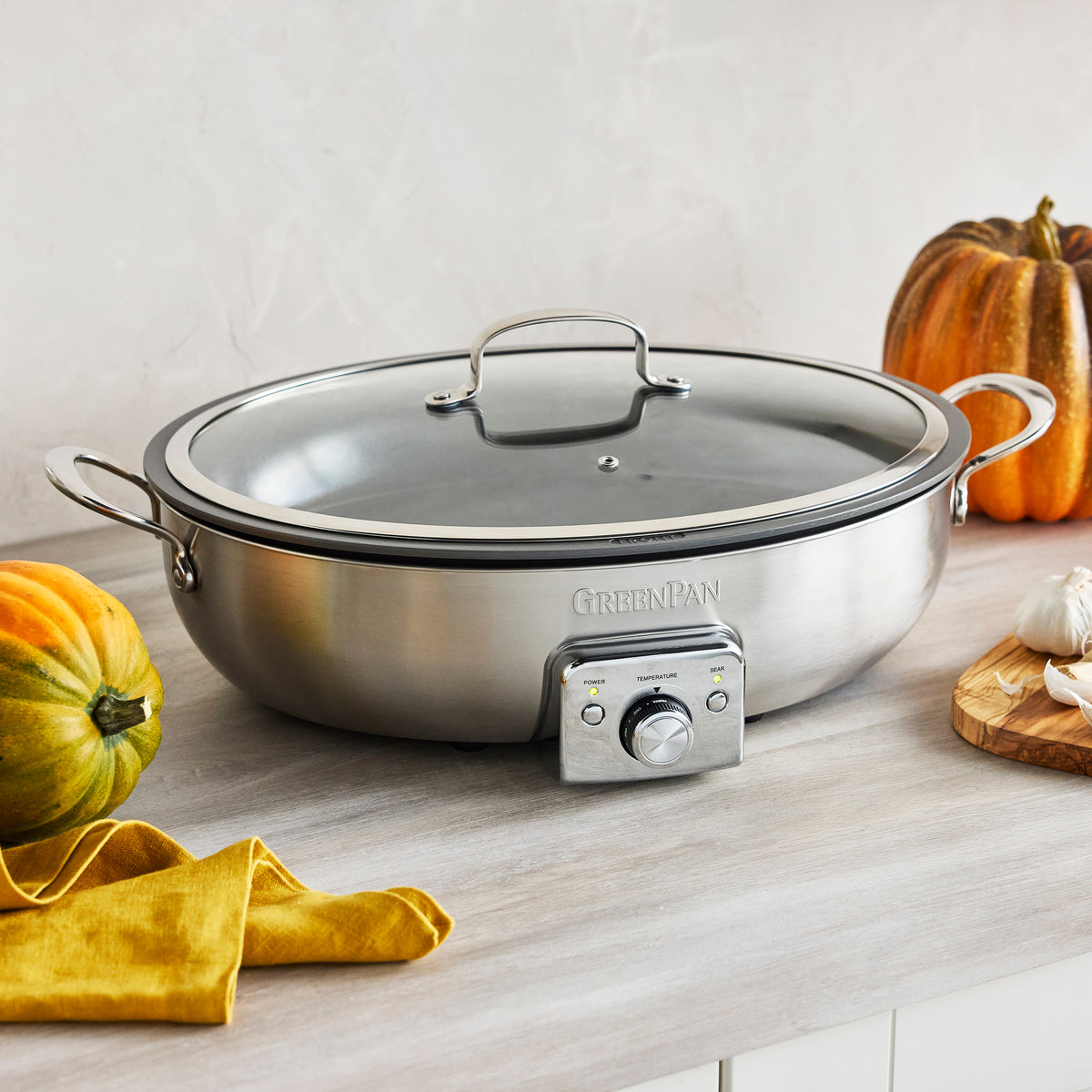 Black oval electric skillet on a kitchen counter next to pumpkins, garlic, and a yellow napkin.