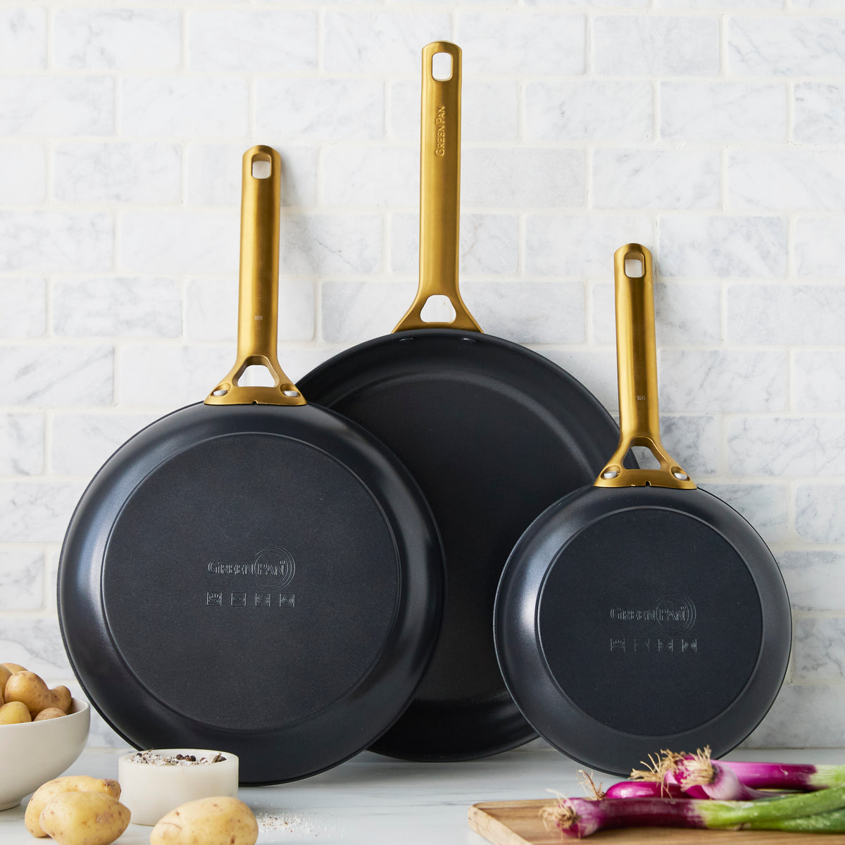 Set of three black frying pans with gold handles on a white tiled wall background