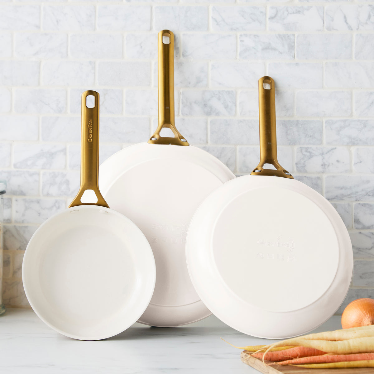 Three cream frying pans with gold handles on a kitchen counter.
