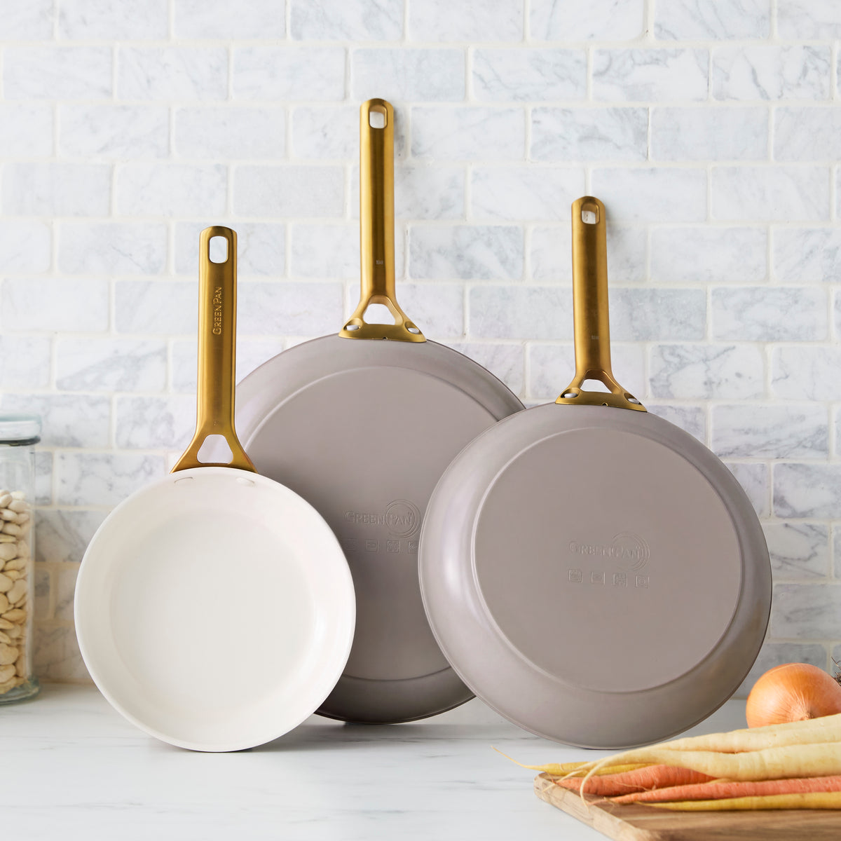 Set of three latte-colored ceramic frypans with gold handles, upright against a white tiled kitchen wall.