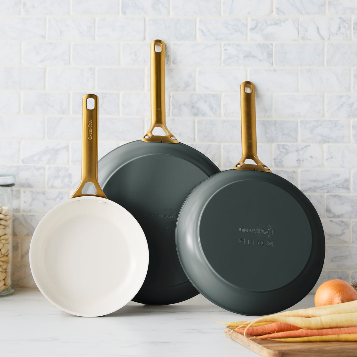 Set of three green ceramic nonstick frypans with gold handles hanging on a white tile wall near fresh vegetables and beans.