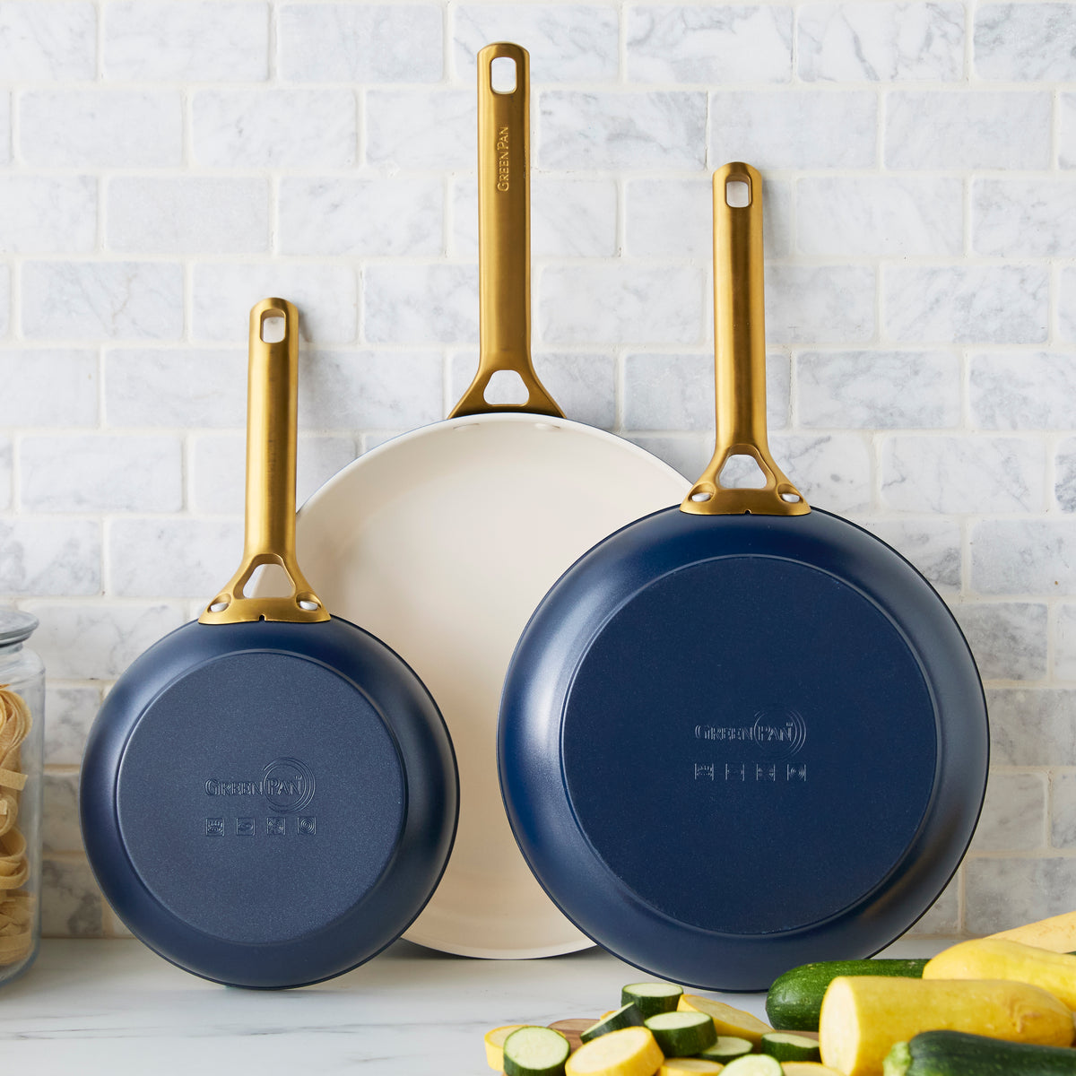 Three dark blue frying pans with gold handles on a kitchen counter with vegetables