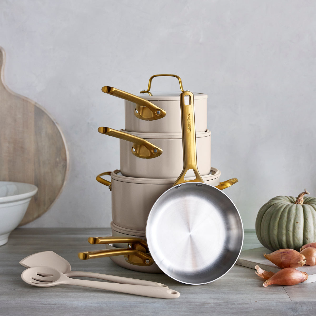 12-piece stainless steel cookware set in macadamia displayed on a counter with utensils, a pumpkin, shallots, and a bowl.