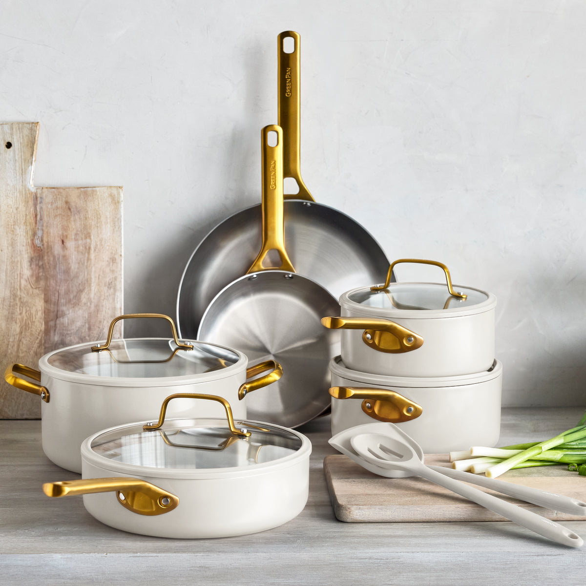 Cream stainless steel 12-piece cookware set with gold handles on a counter, next to green onions and kitchen utensils.