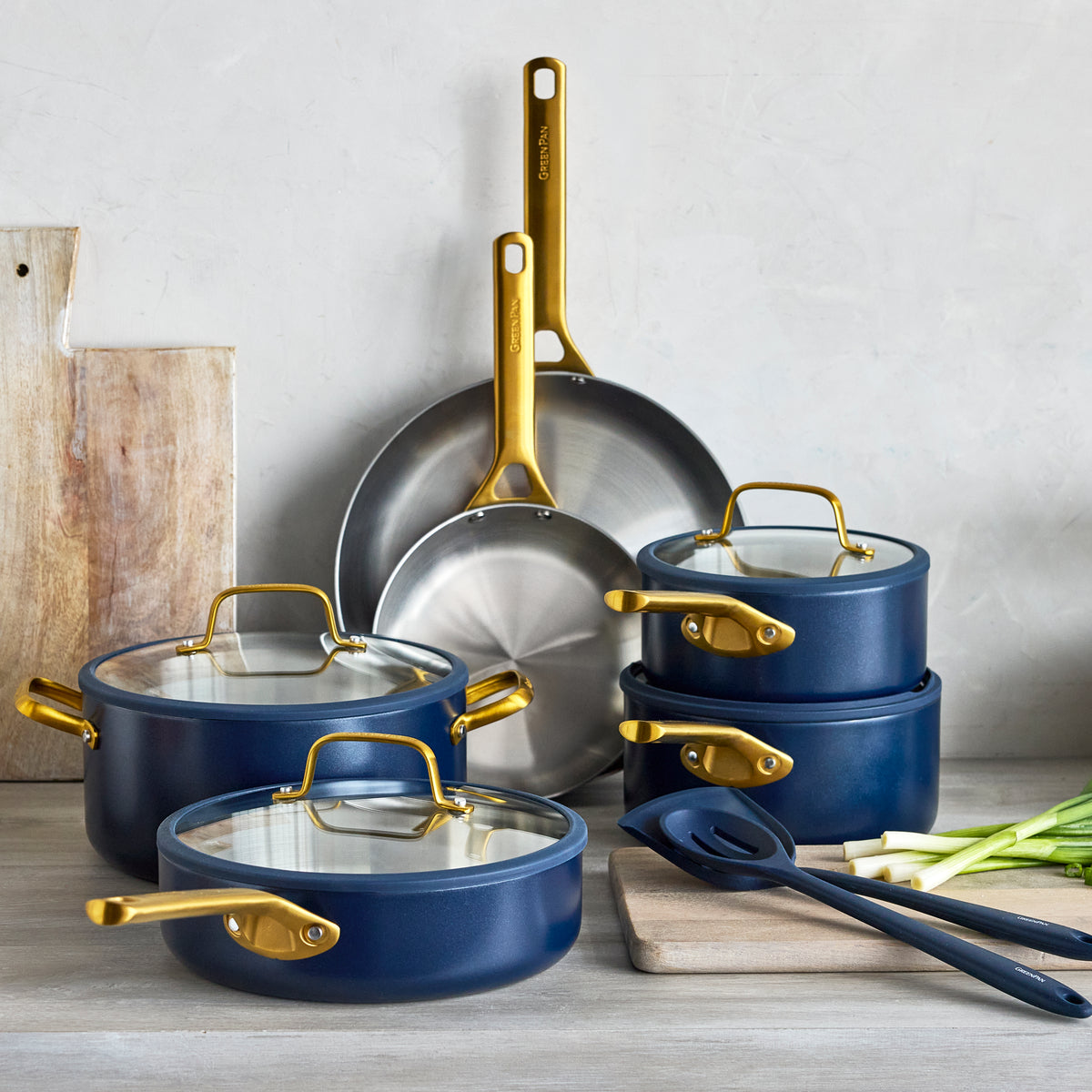 Stainless steel 12-piece cookware set in twilight color on a kitchen counter with cutting boards, utensils, and green onions nearby.